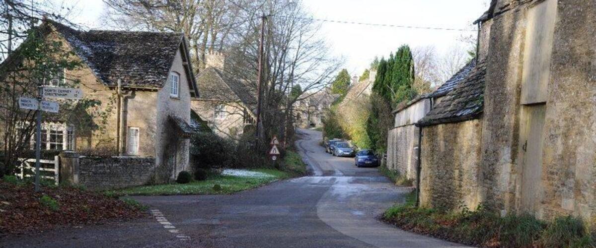 A narrow rural road flanked by stone buildings, a signpost, and a pedestrian crossing.