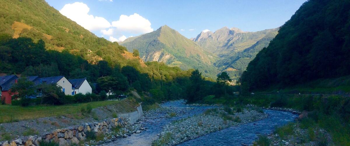 A river flowing through a valley with houses on the left, surrounded by mountains.