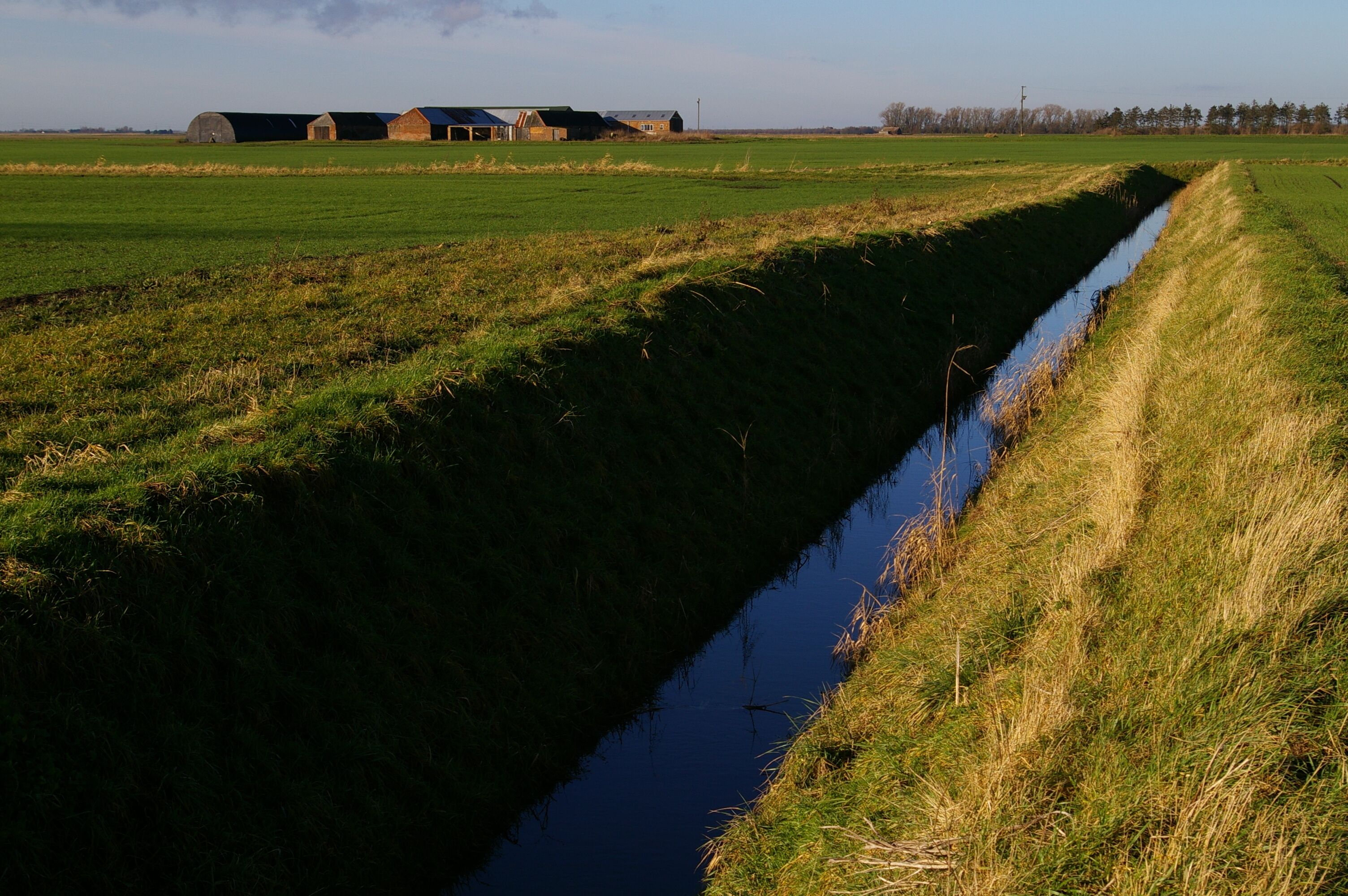 A narrow water channel with grass on either side, a barn, and open fields in the background.