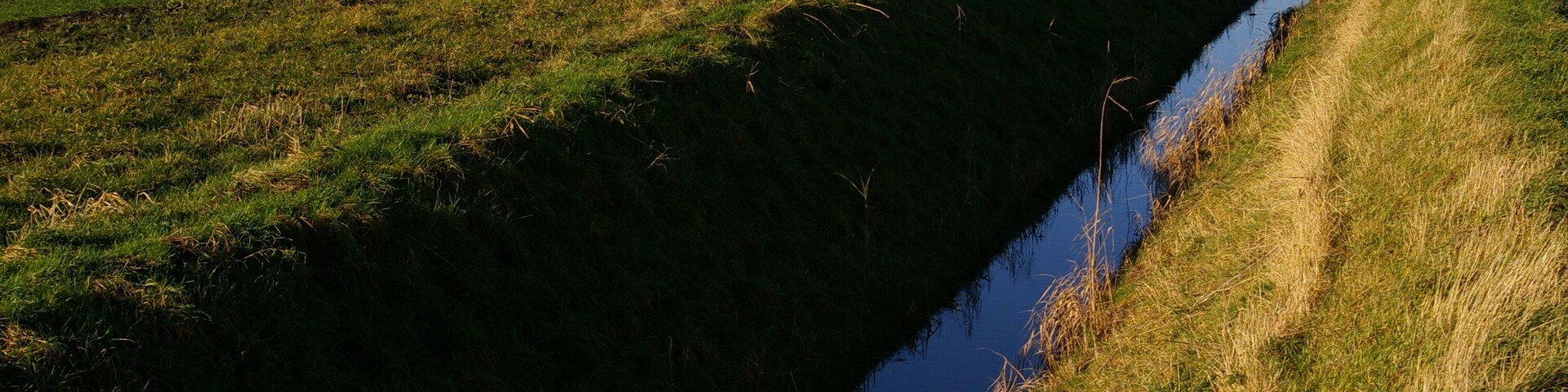 A narrow water channel with grass on either side, a barn, and open fields in the background.