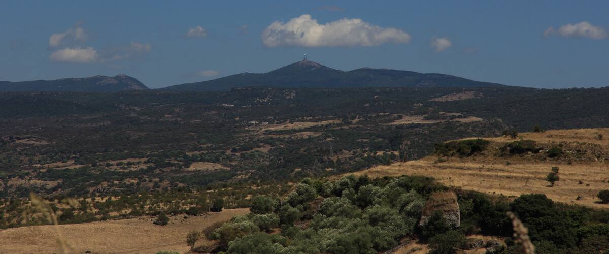 A mountainous landscape with a prominent peak, surrounded by dry grasslands and scattered trees.