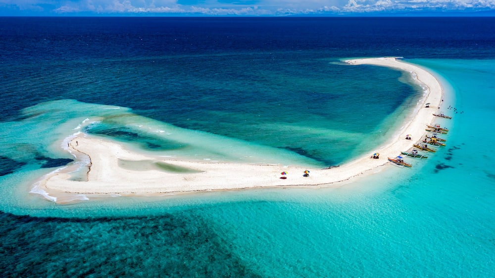 Eine Vogelperspektive zeigt einen sichelförmigen, weißen Sandstrand mit klarem, türkisblauem Wasser, umgeben von Booten und Sonnenschirmen.