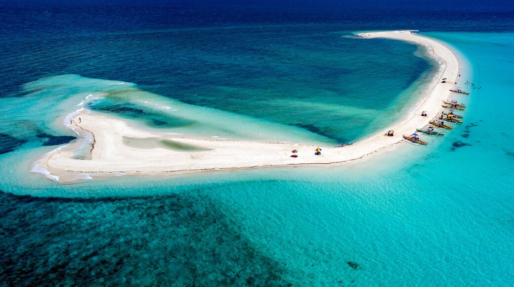 Vista aérea de una playa de arena blanca en forma de media luna, con aguas turquesas cristalinas, rodeada de botes y sombrillas.