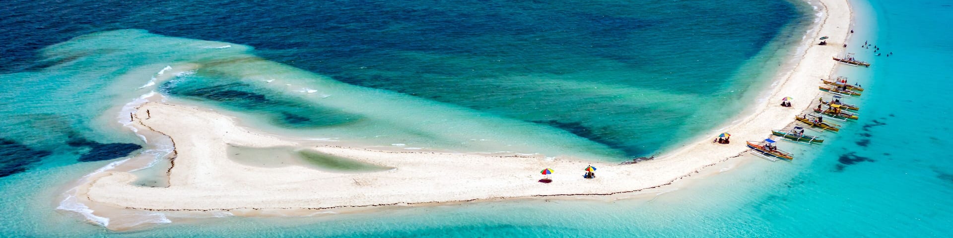 An aerial view of a crescent-shaped white sand beach with clear turquoise waters, surrounded by boats and umbrellas.
