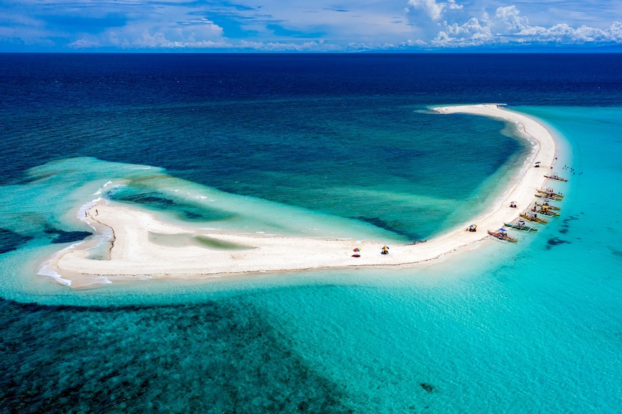 Vista aérea de una playa de arena blanca en forma de media luna, con aguas turquesas cristalinas, rodeada de botes y sombrillas.