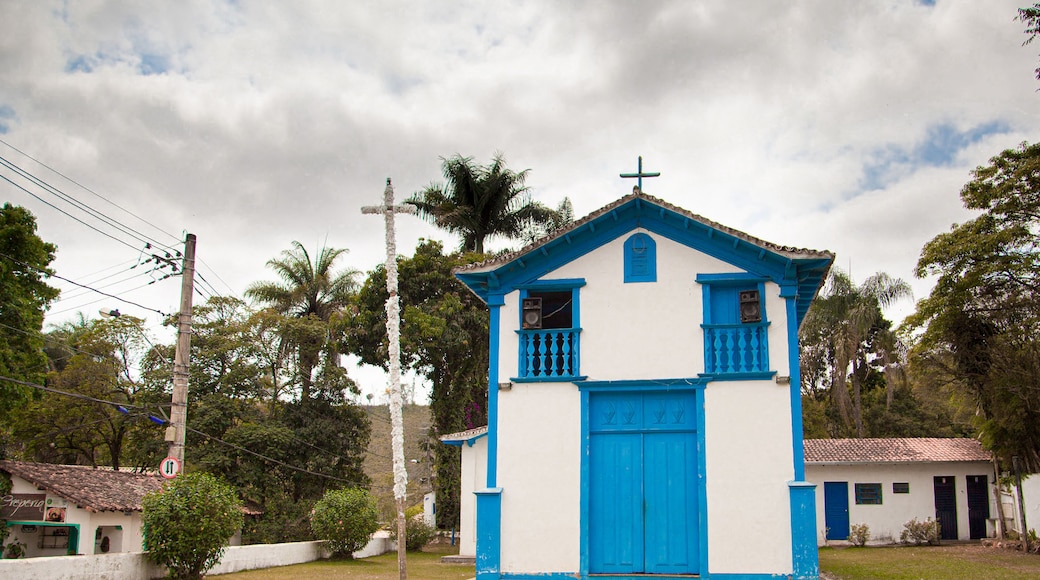 A small church with a blue door and white walls, surrounded by greenery and a cloudy sky.