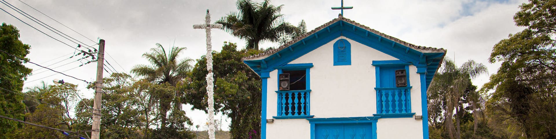 A small church with a blue door and white walls, surrounded by greenery and a cloudy sky.
