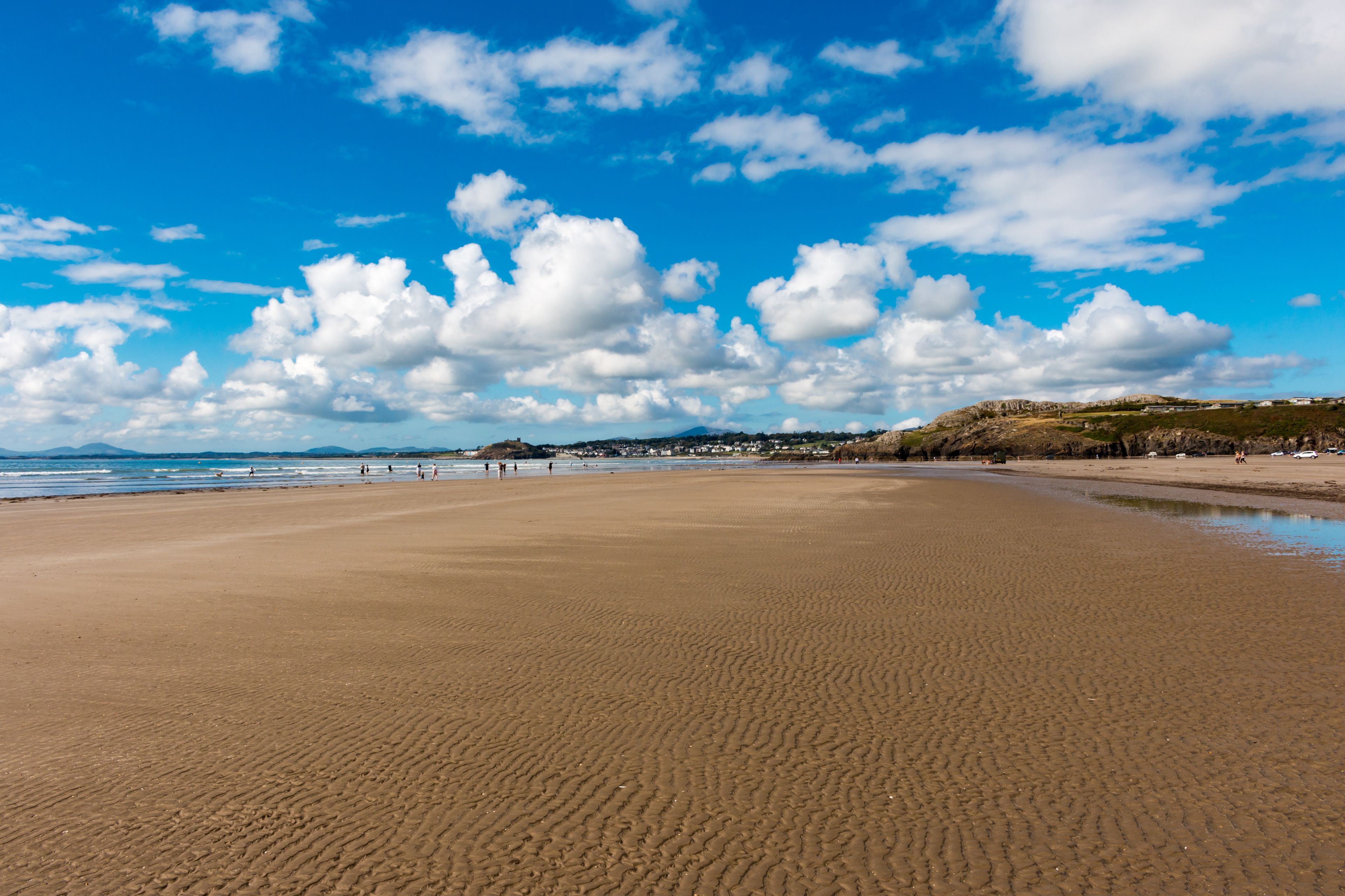 A wide sandy beach with gentle waves, a clear blue sky, and scattered clouds.