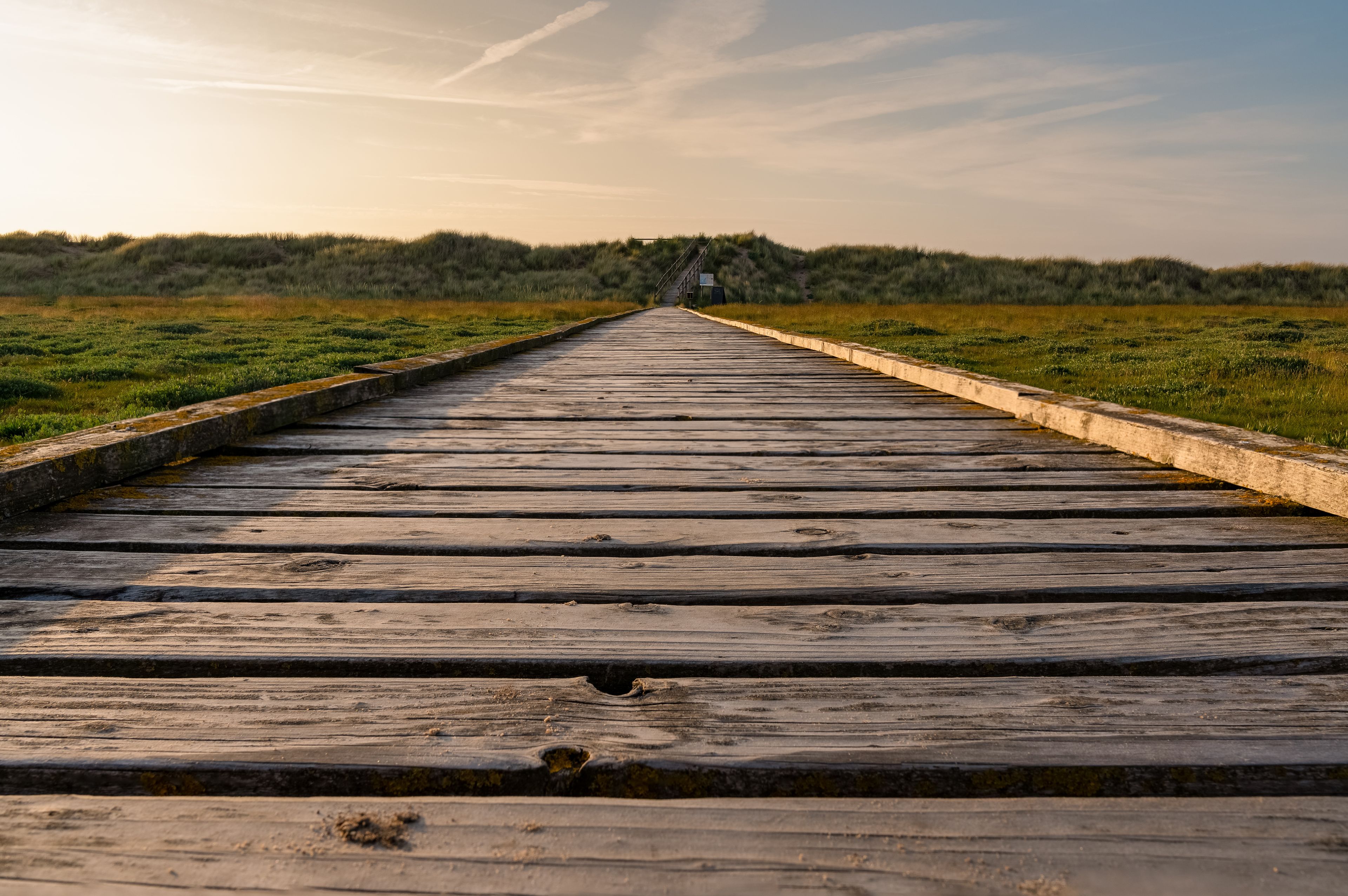 A wooden boardwalk extending into a grassy field with a hill in the distance.