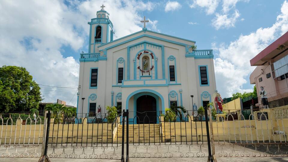 A church with a blue and white facade, a bell tower, and a statue in front.