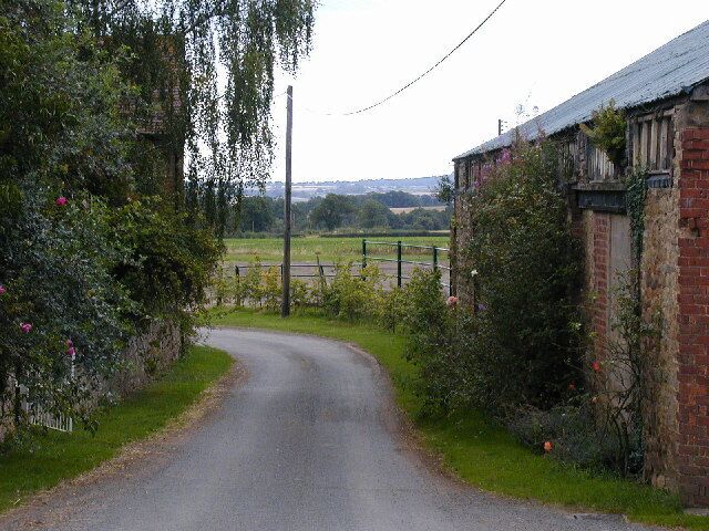 Un camino rural estrecho bordeado por edificios de ladrillo y abundante vegetación.