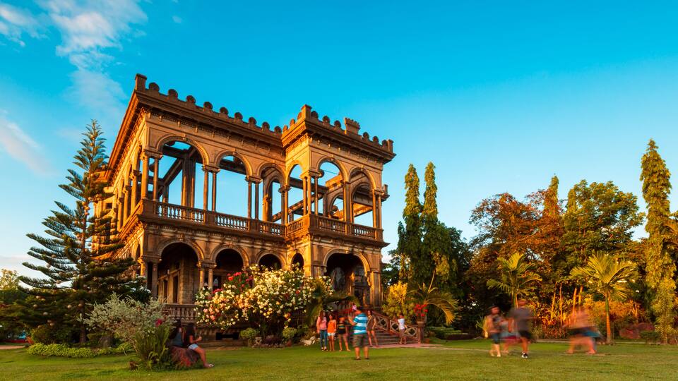 A historical building with arches and columns, surrounded by a well-maintained garden with trees and flowers.