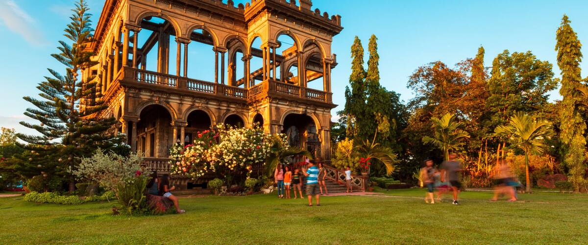 A historical building with arches and columns, surrounded by a well-maintained garden with trees and flowers.