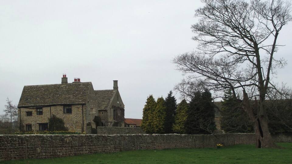 A stone house with a sloped roof and chimneys, surrounded by a stone wall and a grassy field.