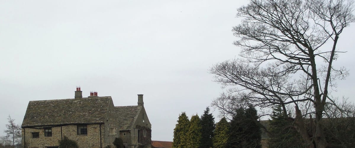 A stone house with a sloped roof and chimneys, surrounded by a stone wall and a grassy field.