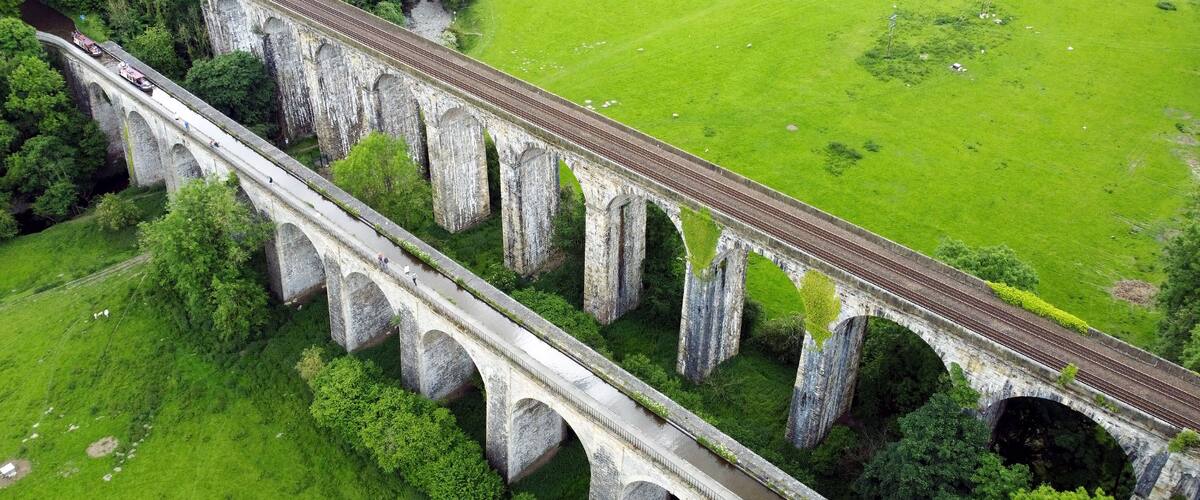 Um longo viaduto de pedra atravessa um rio, rodeado por campos verdes e árvores.