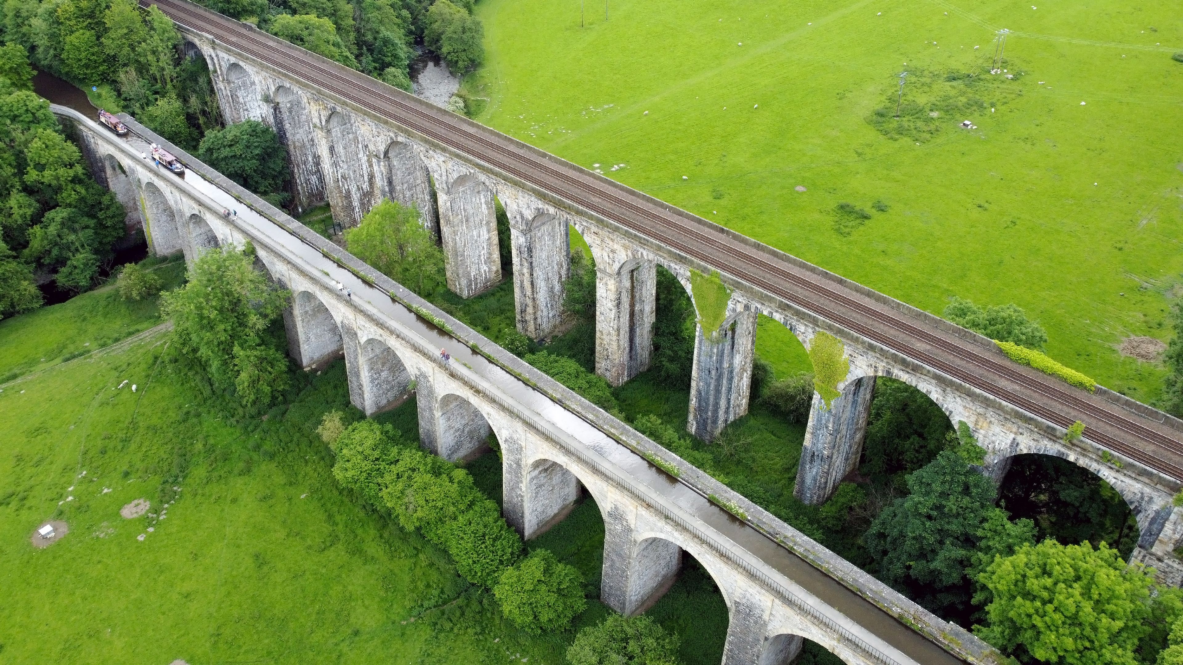 Vista aérea de un viaducto de piedra con múltiples arcos que cruzan un río, rodeado de exuberantes campos verdes y árboles.
