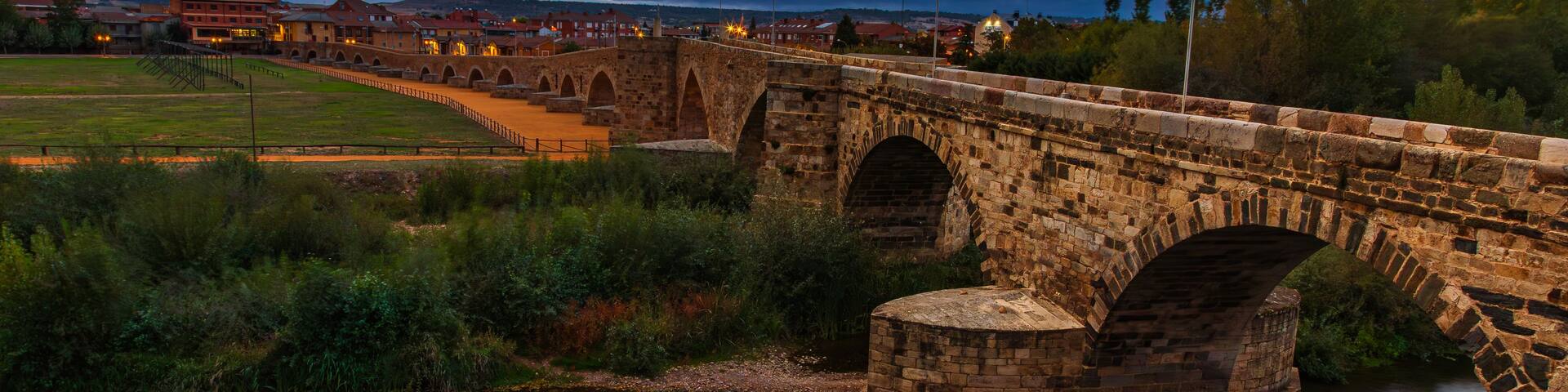 A stone bridge over a river with a town in the background under a cloudy sky.