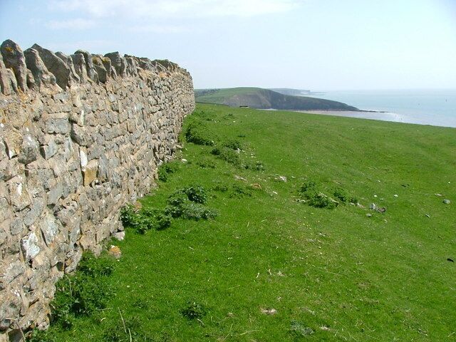 Mural de pedra, campo com relva e costa marítima.
