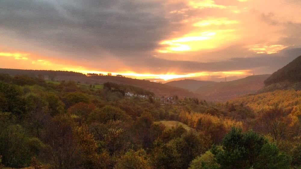 A valley with autumn foliage, a river, and distant hills under a sunset sky.