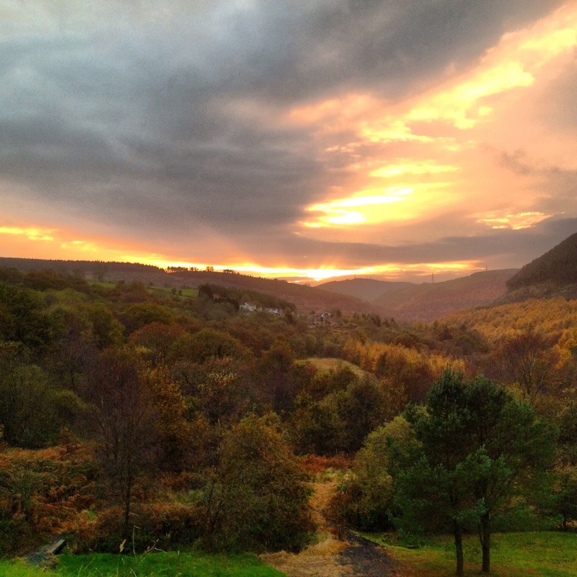 A valley with autumn foliage, a river, and distant hills under a sunset sky.