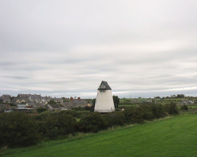 Ein weißer Leuchtturm mit einer kegelförmigen Spitze, umgeben von Grün und Häusern im Hintergrund.