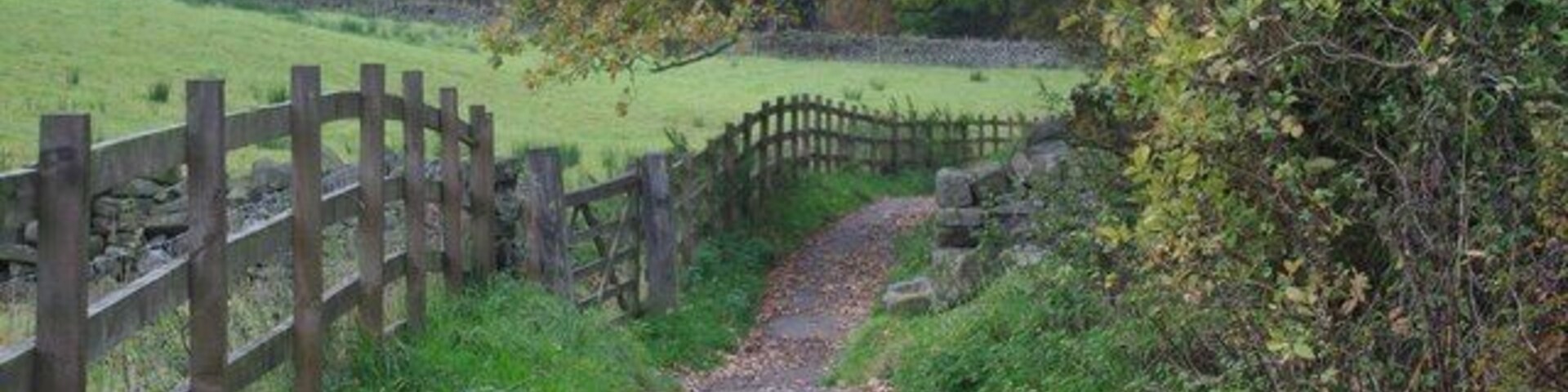 Un sendero de grava estrecho flanqueado por una valla de madera a un lado y un muro de piedra al otro, que atraviesa un campo verde.