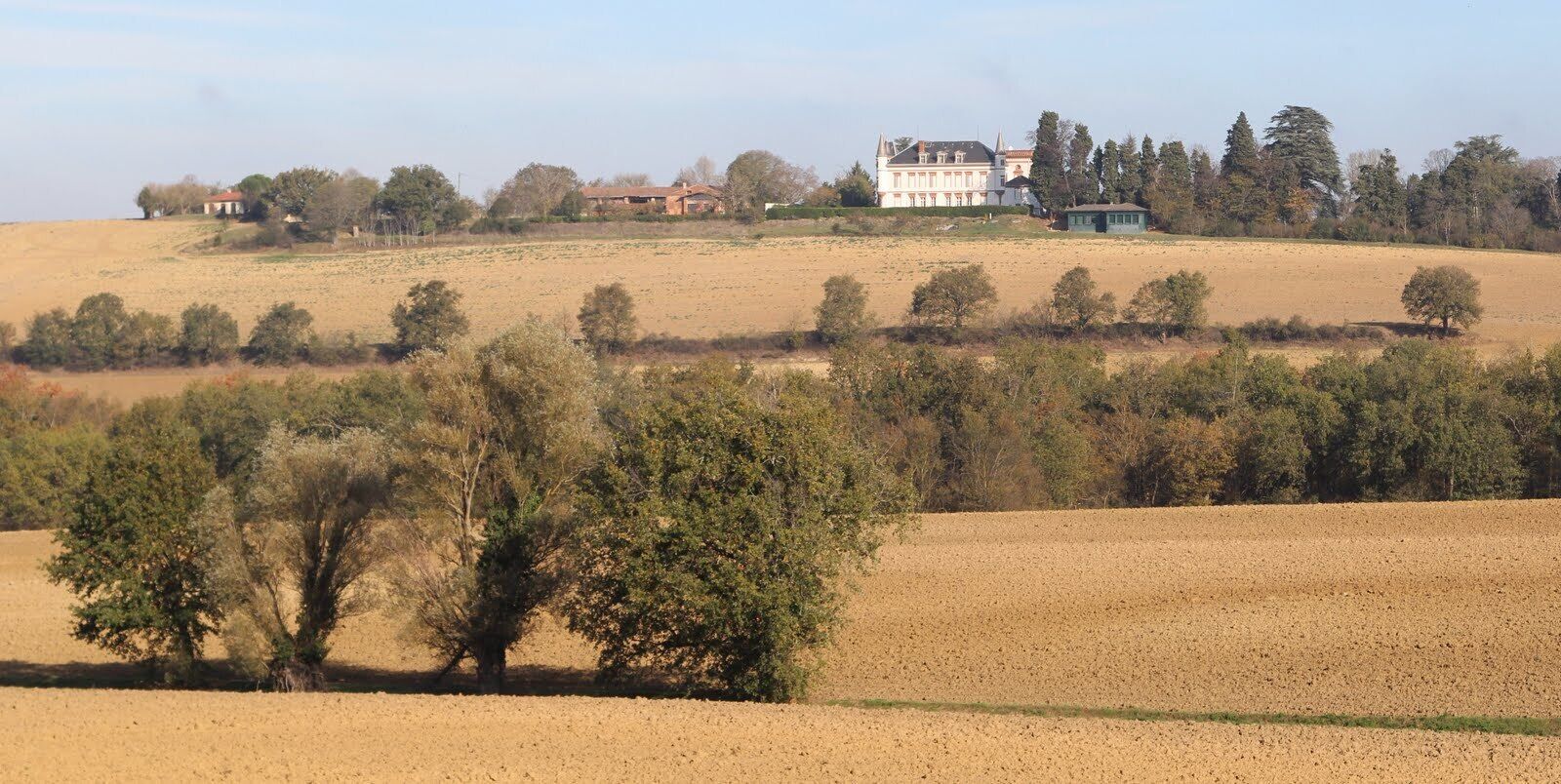 A rural landscape with rolling hills, scattered trees, and a large white house with a red roof on a hill.