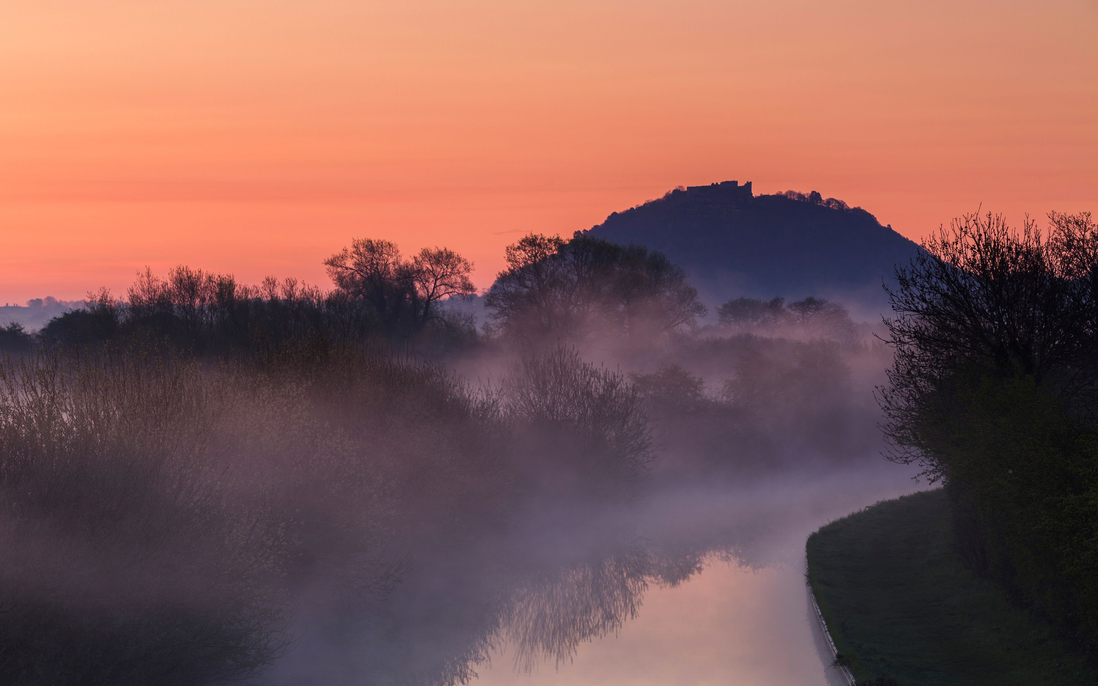 A misty landscape with a river, trees, and a hill during sunrise or sunset.