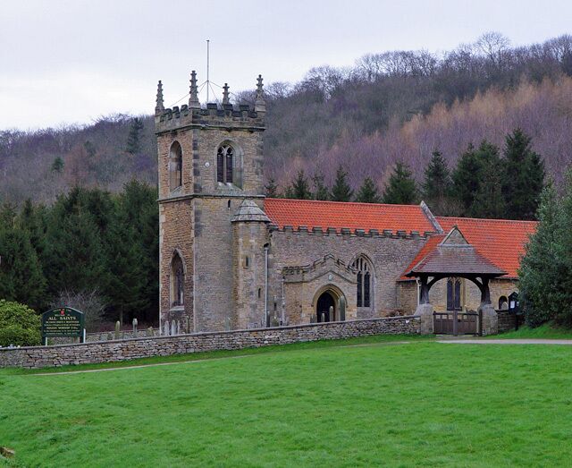 A historic stone church with a red-tiled roof and a bell tower, surrounded by a grassy field and dense forest.