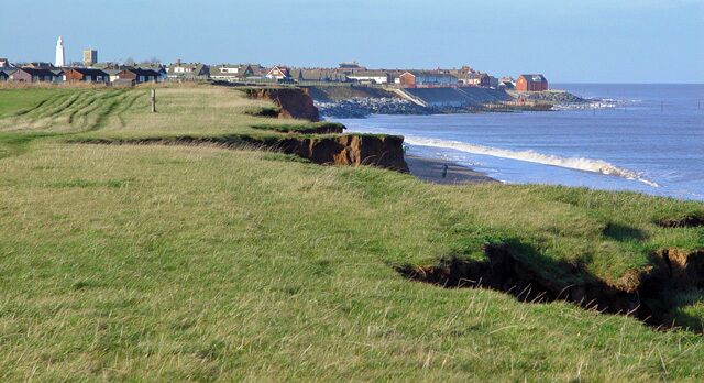 A coastal landscape with grassy fields, cliffs, and a lighthouse in the distance.