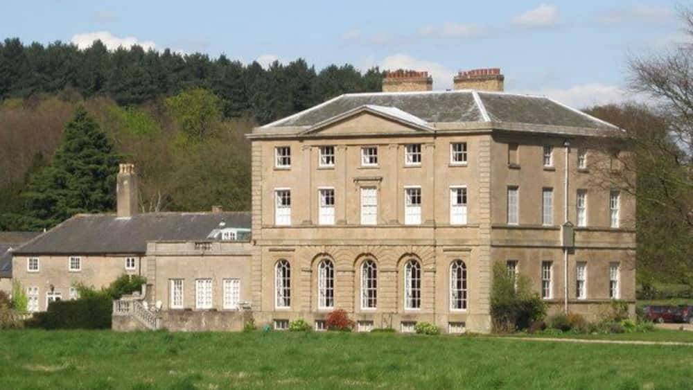 Un grand bâtiment historique en pierre, doté de nombreuses fenêtres et d’une façade symétrique, entouré d’un champ verdoyant et d’arbres.