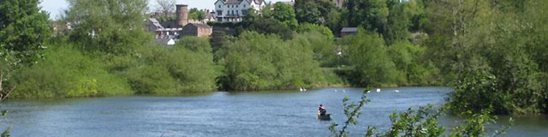 A river with a person rowing, surrounded by greenery and a church with a spire in the background.