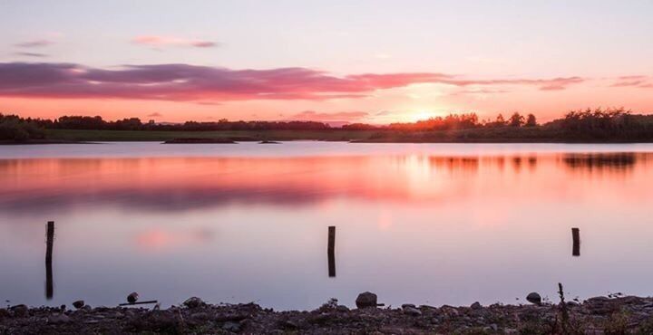 A calm lake at sunset with a reflection of the sky, surrounded by a shoreline with rocks and a distant tree line.