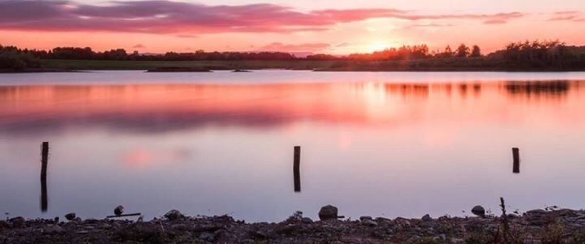 A calm lake at sunset with a reflection of the sky, surrounded by a shoreline with rocks and a distant tree line.