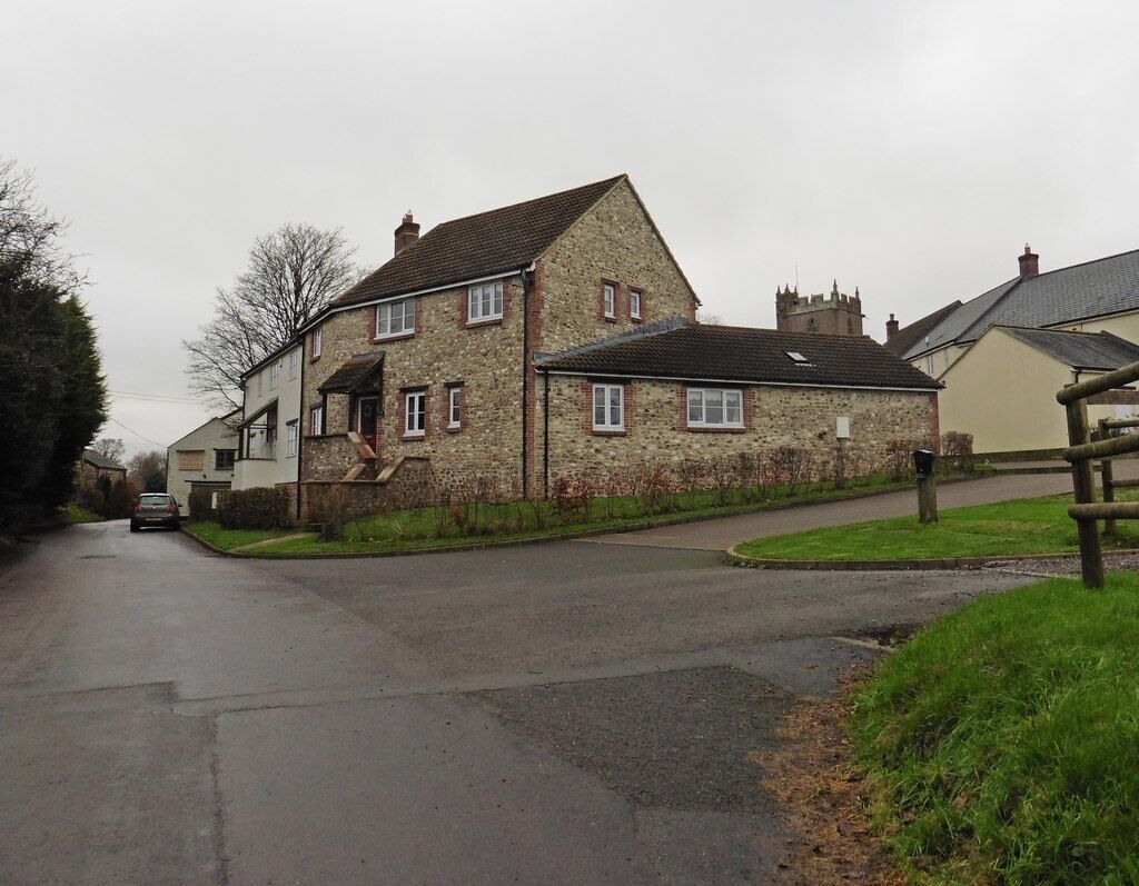 A stone house with a sloped roof, a paved driveway, and a grassy area with a wooden fence.