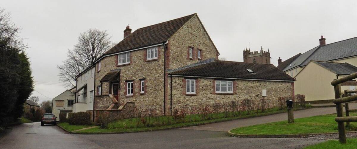 A stone house with a sloped roof, a paved driveway, and a grassy area with a wooden fence.