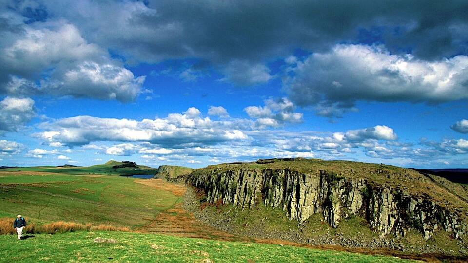 A grassy field with a rocky cliff and a blue sky with clouds.