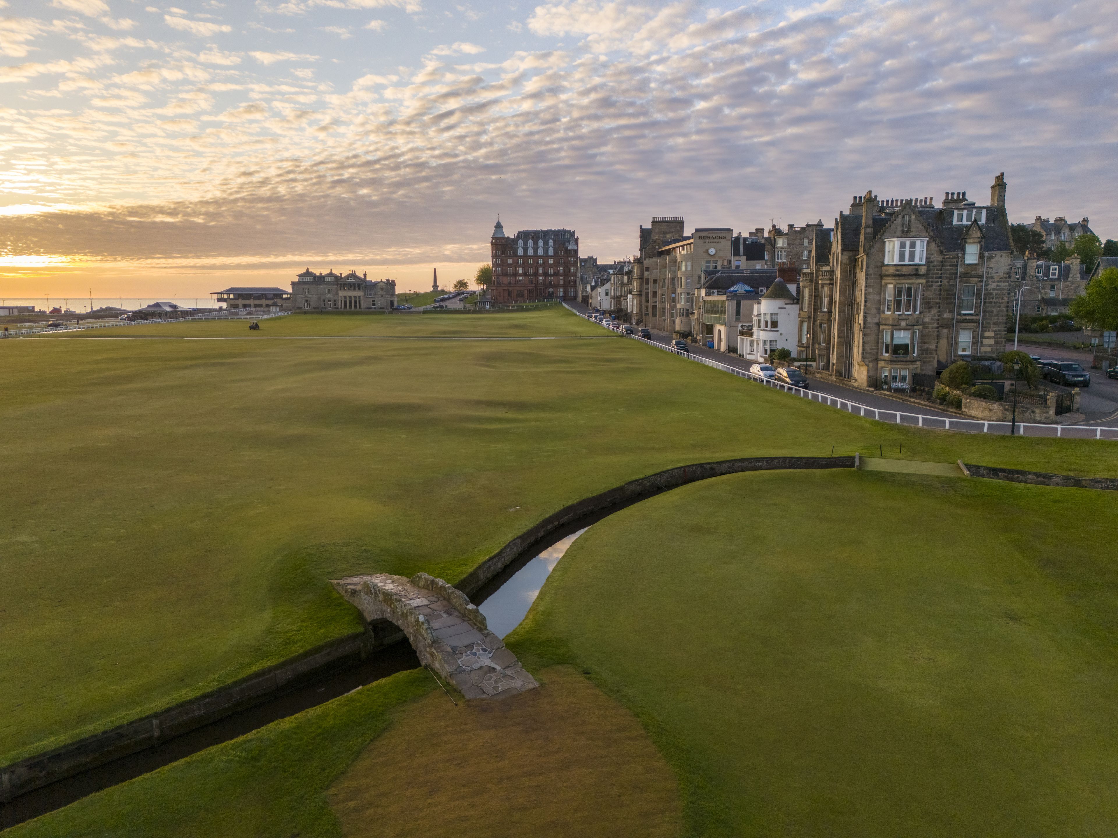 Un campo de golf con un puente de piedra, un edificio histórico y una hilera de casas residenciales.