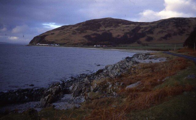 A coastal landscape with a rocky shoreline, a grassy hill, and a road leading to a small settlement by the sea.