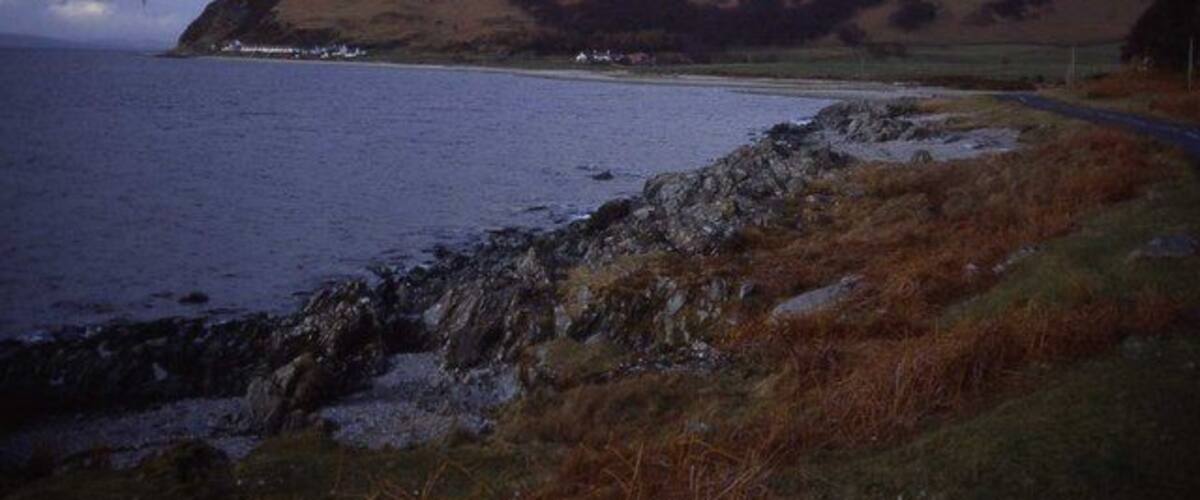 A coastal landscape with a rocky shoreline, a grassy hill, and a road leading to a small settlement by the sea.