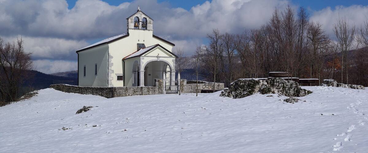 A white church with a bell tower and arched entrance, surrounded by snow-covered landscape and trees.