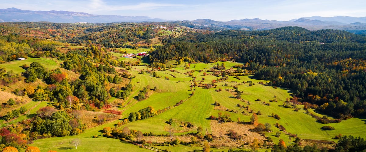 A panoramic view of a mountainous landscape with rolling green fields, dense forests, and distant mountain ranges under a clear blue sky.