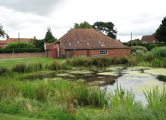 View across duck pond. The pond is located on the village green, beside the B1149 (Norwich Road) > 877231. The brick building seen beyond is the village hall.