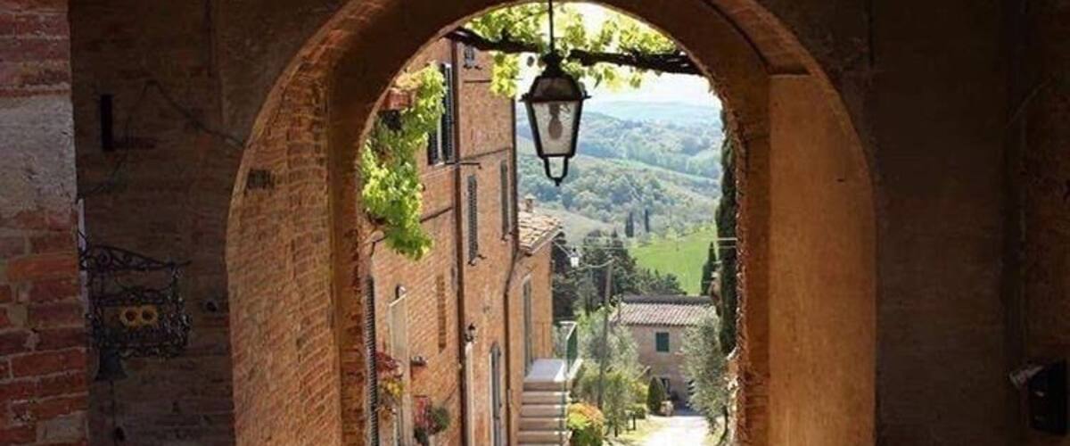 An arched stone gateway leading to a cobblestone street with buildings and a view of rolling hills in the distance.
