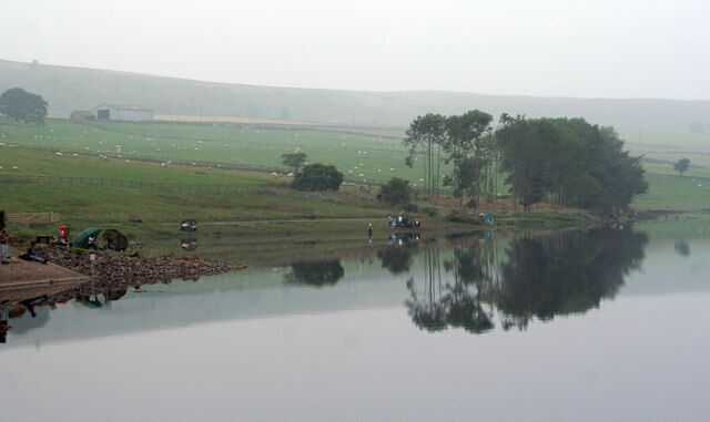 A calm lake with a reflection of trees and a distant farm.