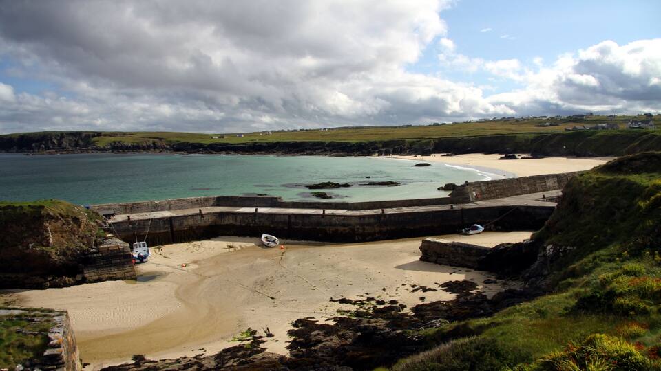 A coastal beach with a rocky breakwater, calm turquoise waters, and a grassy hillside.