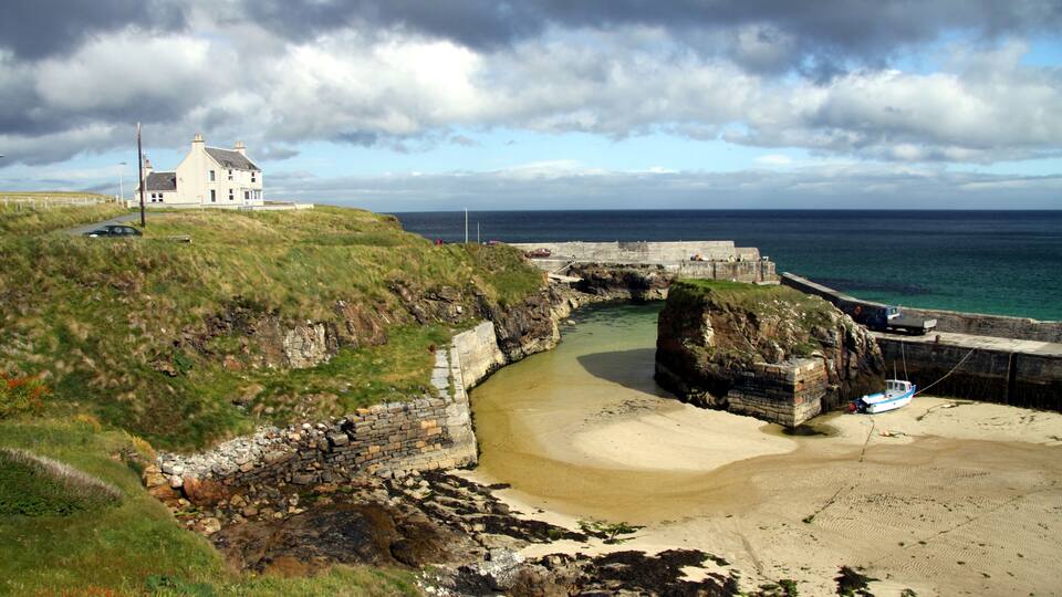 A coastal landscape with a white house, a stone bridge, and a sandy beach.