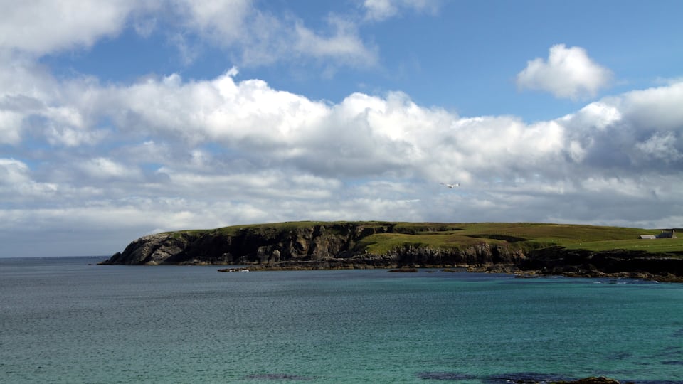 A coastal landscape with a grassy hill, rocky shoreline, and a clear blue sky with clouds.