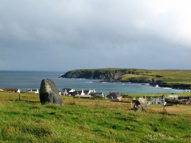 A coastal landscape with a prominent standing stone, grassy fields, and a small settlement with white-roofed buildings by the sea.
